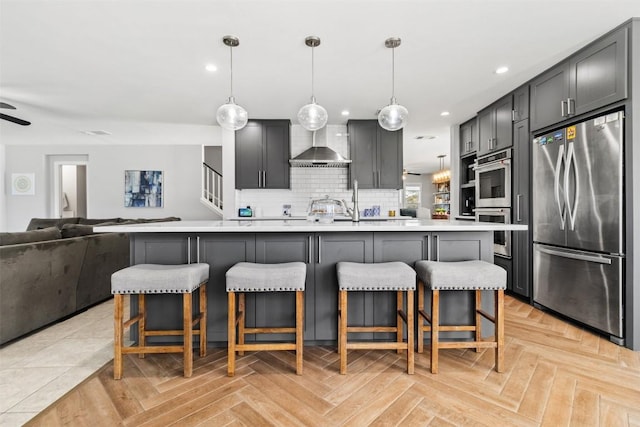 kitchen with stainless steel appliances, wall chimney range hood, decorative light fixtures, a center island with sink, and light parquet flooring