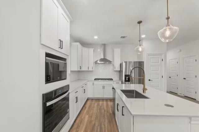 kitchen with wall chimney exhaust hood, stainless steel appliances, pendant lighting, a center island with sink, and white cabinets