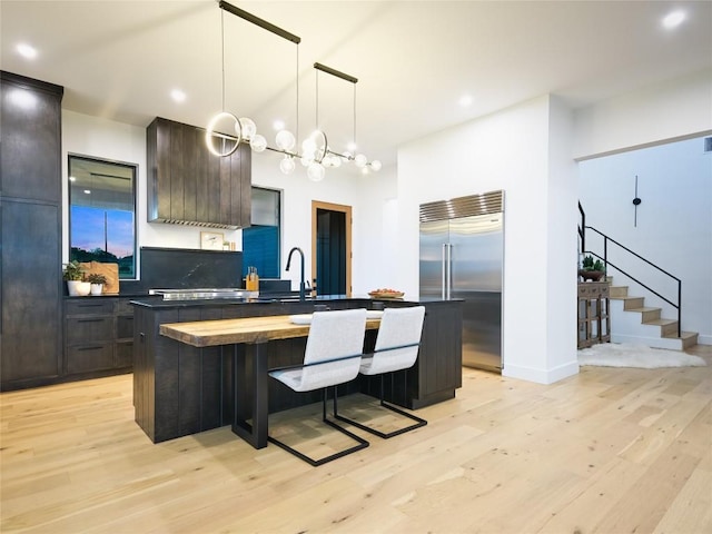 kitchen featuring light wood-type flooring, tasteful backsplash, dark brown cabinetry, a kitchen island with sink, and built in fridge