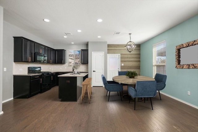 kitchen with dark hardwood / wood-style flooring, backsplash, black appliances, decorative light fixtures, and an island with sink