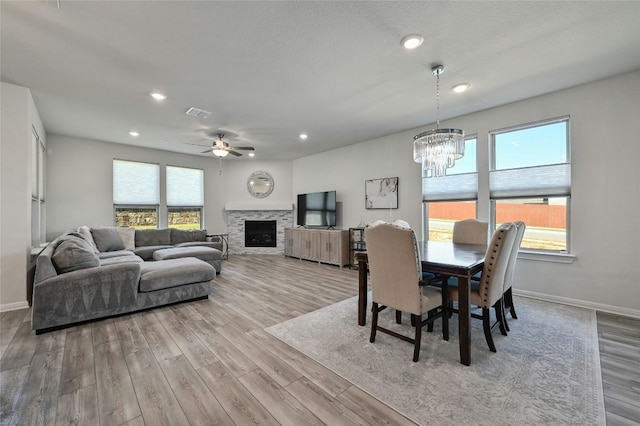 dining area with a stone fireplace, ceiling fan with notable chandelier, and light wood-type flooring