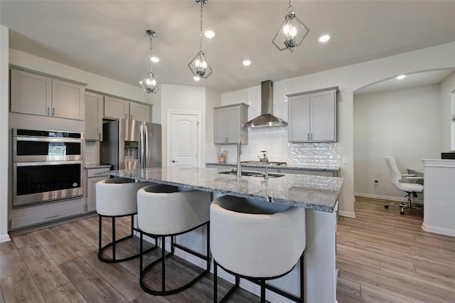 kitchen with appliances with stainless steel finishes, a kitchen island with sink, light stone countertops, decorative light fixtures, and wall chimney exhaust hood