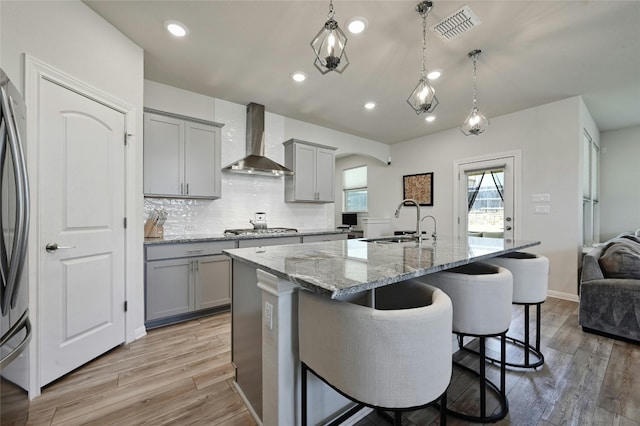 kitchen featuring gray cabinets, pendant lighting, sink, a kitchen island with sink, and wall chimney exhaust hood