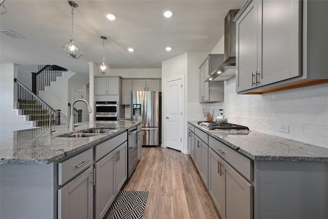 kitchen featuring sink, a center island with sink, appliances with stainless steel finishes, pendant lighting, and wall chimney range hood