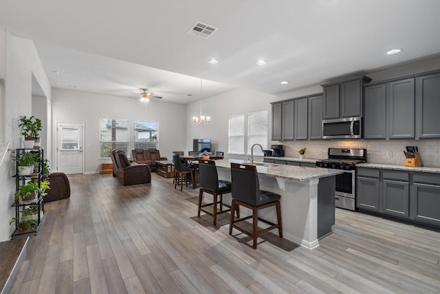 kitchen featuring gray cabinetry, ceiling fan with notable chandelier, an island with sink, light stone counters, and stainless steel appliances