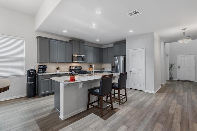 kitchen with gray cabinetry, a kitchen island with sink, hanging light fixtures, light stone countertops, and stainless steel appliances