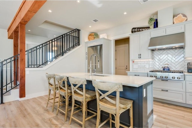 kitchen with ventilation hood, sink, light hardwood / wood-style flooring, white cabinetry, and an island with sink