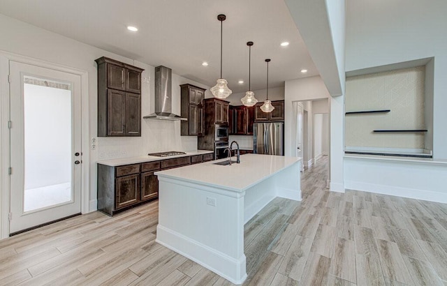 kitchen featuring wall chimney range hood, sink, an island with sink, decorative light fixtures, and stainless steel appliances