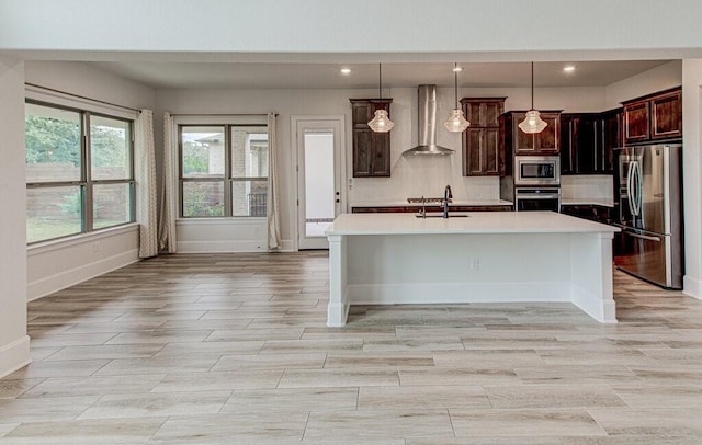 kitchen with stainless steel appliances, sink, wall chimney range hood, hanging light fixtures, and an island with sink