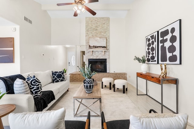 living room featuring a towering ceiling, light wood-type flooring, ceiling fan, beam ceiling, and a stone fireplace