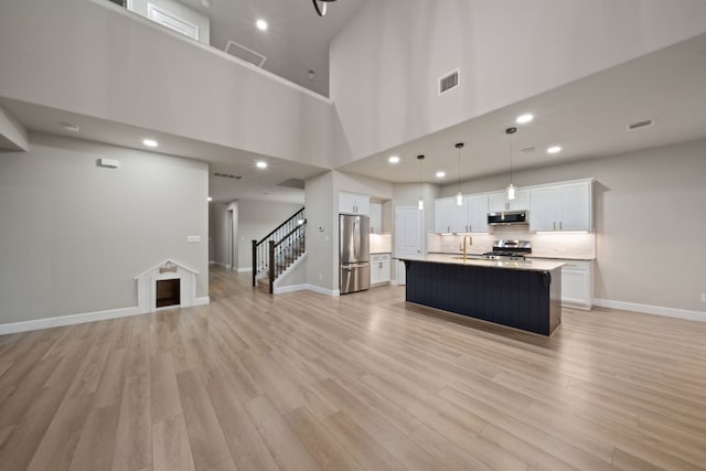 kitchen featuring white cabinetry, stainless steel appliances, a towering ceiling, light hardwood / wood-style floors, and a center island with sink