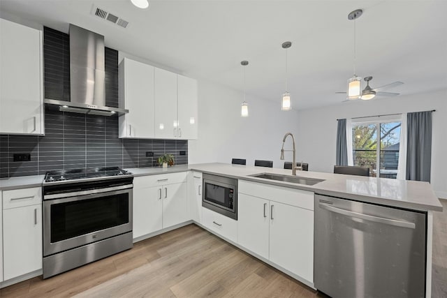 kitchen featuring sink, wall chimney exhaust hood, kitchen peninsula, white cabinets, and appliances with stainless steel finishes