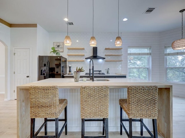 kitchen featuring wall chimney exhaust hood, black refrigerator with ice dispenser, backsplash, a kitchen island with sink, and a breakfast bar