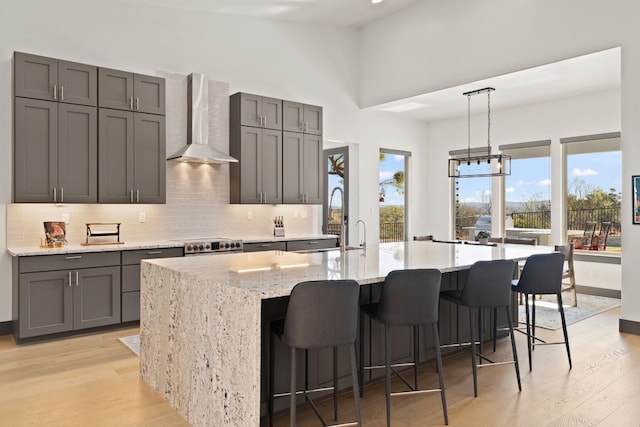 kitchen with gray cabinetry, wall chimney exhaust hood, hanging light fixtures, light hardwood / wood-style flooring, and a large island with sink