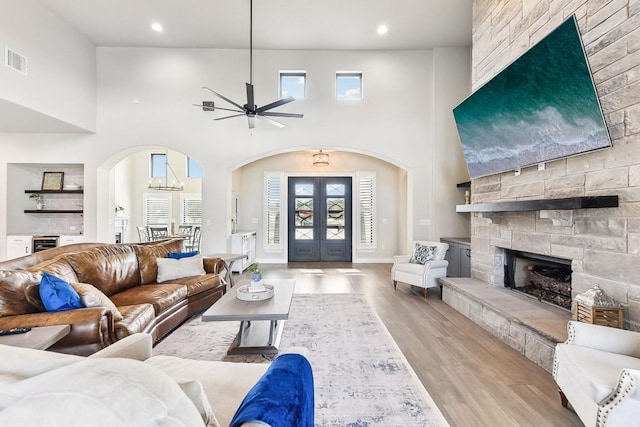 living room with french doors, a high ceiling, a stone fireplace, light hardwood / wood-style flooring, and ceiling fan with notable chandelier