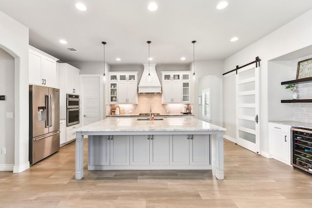 kitchen with a barn door, appliances with stainless steel finishes, an island with sink, white cabinets, and custom exhaust hood