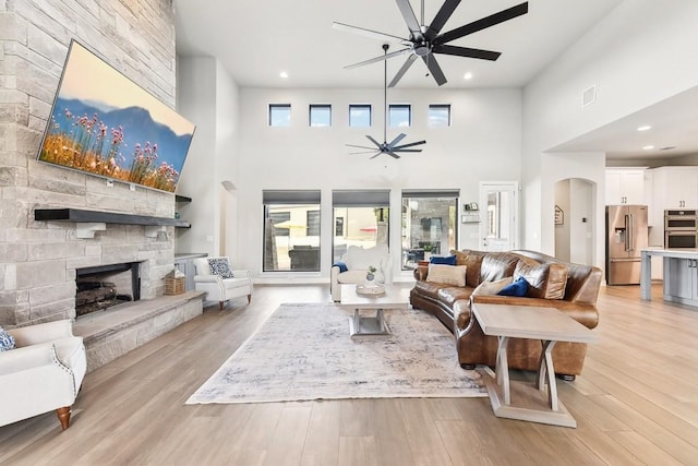 living room featuring a high ceiling, light hardwood / wood-style flooring, ceiling fan, and a stone fireplace