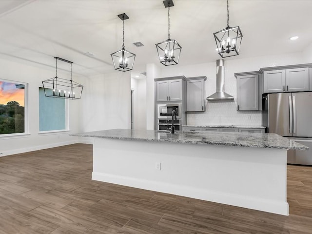 kitchen featuring wall chimney exhaust hood, light stone counters, decorative light fixtures, a kitchen island with sink, and appliances with stainless steel finishes