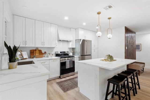 kitchen with light wood-type flooring, custom range hood, stainless steel appliances, decorative light fixtures, and white cabinets