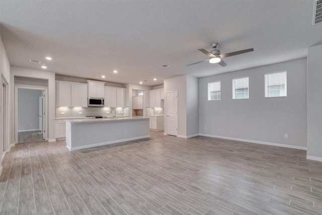 kitchen with white cabinetry, an island with sink, backsplash, ceiling fan, and light wood-type flooring