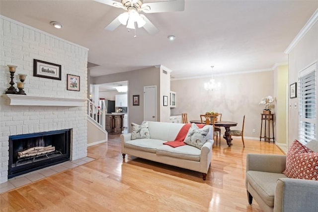 living room featuring a brick fireplace, baseboards, ornamental molding, light wood-style flooring, and ceiling fan with notable chandelier