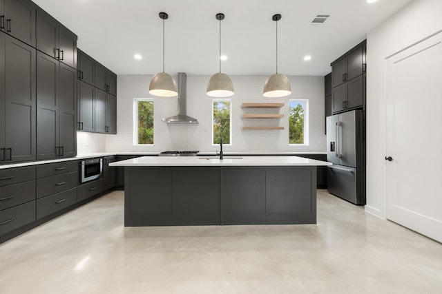 kitchen with sink, wall chimney range hood, an island with sink, decorative light fixtures, and appliances with stainless steel finishes