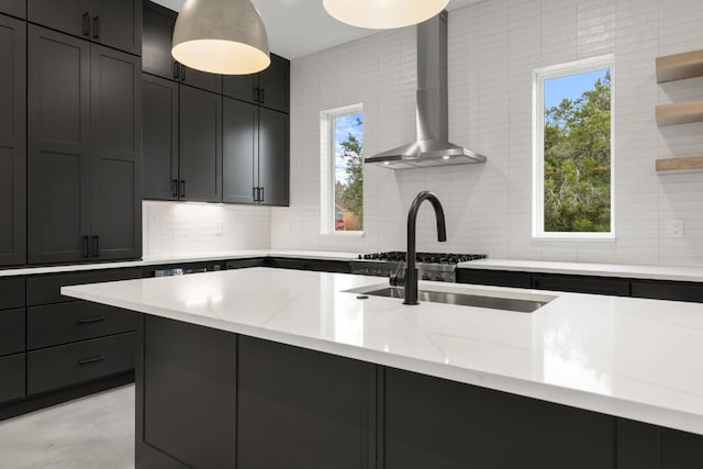 kitchen with light stone countertops, sink, wall chimney exhaust hood, tasteful backsplash, and a kitchen island with sink