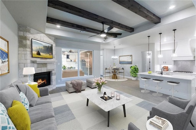 living room featuring sink, light hardwood / wood-style flooring, ceiling fan, beam ceiling, and a stone fireplace