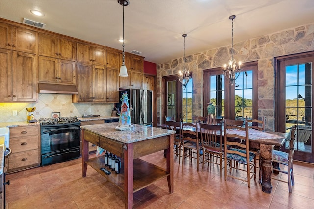 kitchen featuring stainless steel refrigerator, backsplash, light stone countertops, black stove, and decorative light fixtures