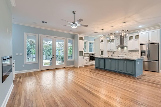 kitchen with appliances with stainless steel finishes, wall chimney range hood, a center island with sink, white cabinets, and light hardwood / wood-style floors