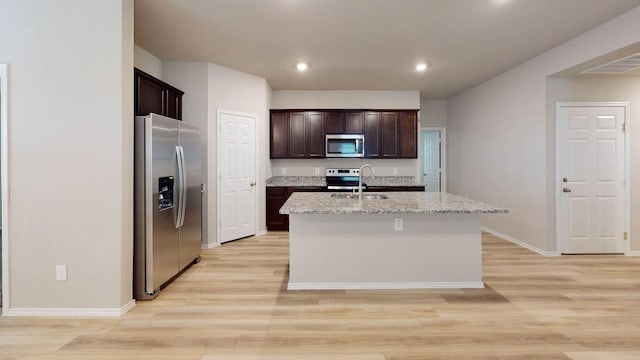 kitchen featuring sink, stainless steel appliances, light stone counters, an island with sink, and light wood-type flooring