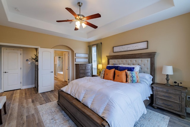 bedroom with a tray ceiling, wood finished floors, arched walkways, baseboards, and ceiling fan