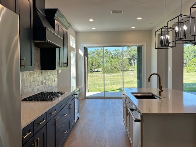 kitchen with appliances with stainless steel finishes, custom range hood, sink, a center island with sink, and hanging light fixtures