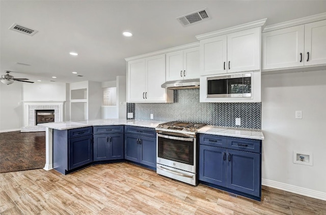 kitchen with appliances with stainless steel finishes, a brick fireplace, blue cabinetry, light hardwood / wood-style flooring, and white cabinets