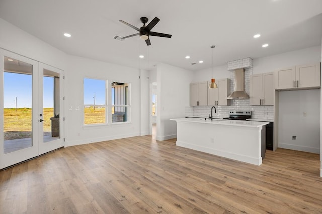 kitchen featuring wall chimney exhaust hood, ceiling fan, decorative light fixtures, light hardwood / wood-style floors, and an island with sink