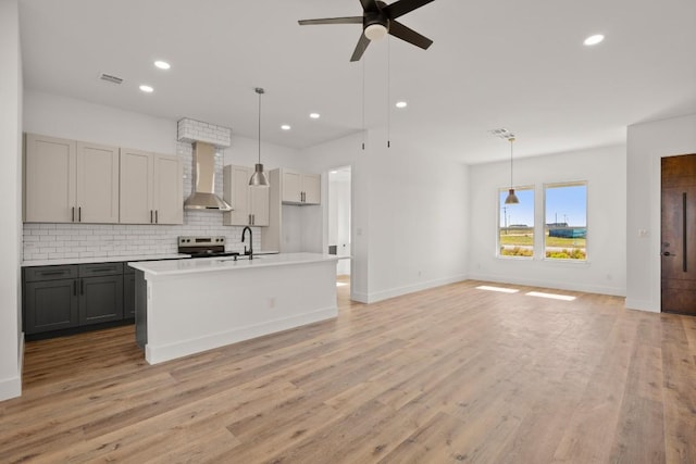 kitchen featuring light wood-type flooring, wall chimney exhaust hood, a kitchen island with sink, gray cabinets, and hanging light fixtures