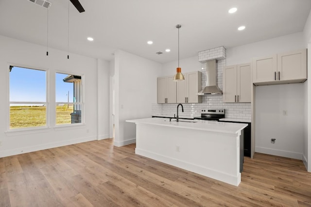 kitchen featuring wall chimney exhaust hood, hanging light fixtures, light hardwood / wood-style flooring, an island with sink, and stainless steel range with electric stovetop