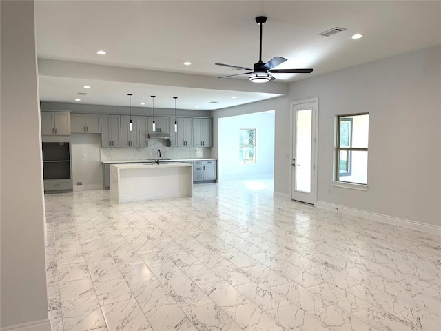 kitchen featuring gray cabinetry, ceiling fan, hanging light fixtures, backsplash, and an island with sink