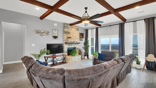 living room featuring beamed ceiling, light hardwood / wood-style floors, a stone fireplace, and ceiling fan