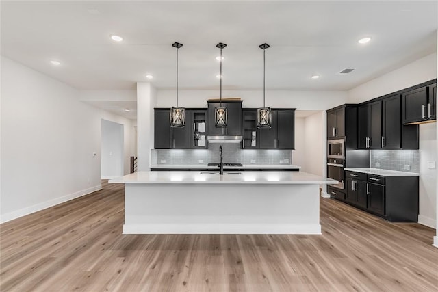 kitchen with hanging light fixtures, light wood-type flooring, stainless steel microwave, black oven, and a kitchen island with sink