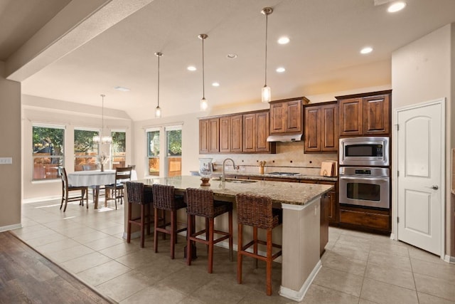 kitchen featuring sink, stainless steel appliances, light stone counters, decorative light fixtures, and a center island with sink