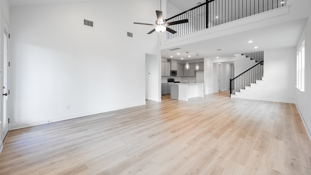 unfurnished living room featuring ceiling fan, a high ceiling, stairs, light wood-style floors, and baseboards