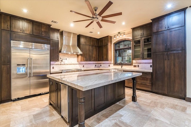 kitchen with wall chimney range hood, dark brown cabinets, light stone countertops, a kitchen island, and built in fridge