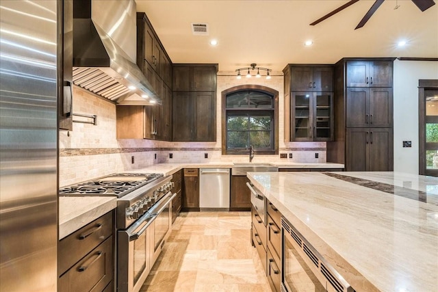 kitchen with stainless steel appliances, tasteful backsplash, light stone counters, dark brown cabinetry, and wall chimney exhaust hood