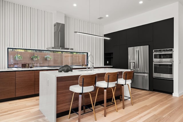 kitchen featuring a center island with sink, wall chimney range hood, light hardwood / wood-style flooring, appliances with stainless steel finishes, and a kitchen bar