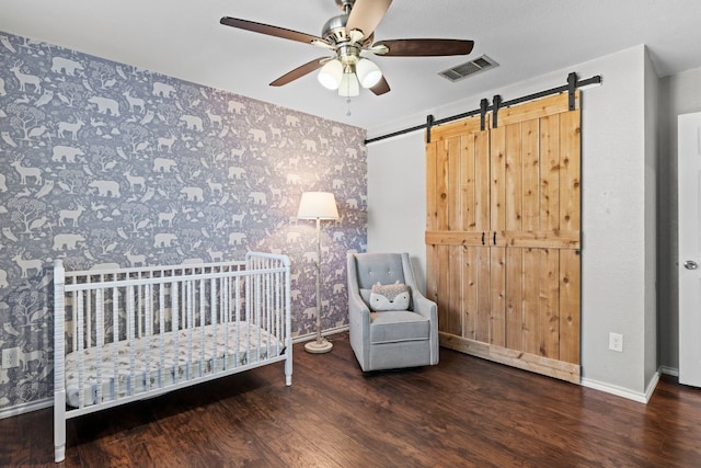 bedroom featuring a crib, dark hardwood / wood-style flooring, a barn door, and ceiling fan