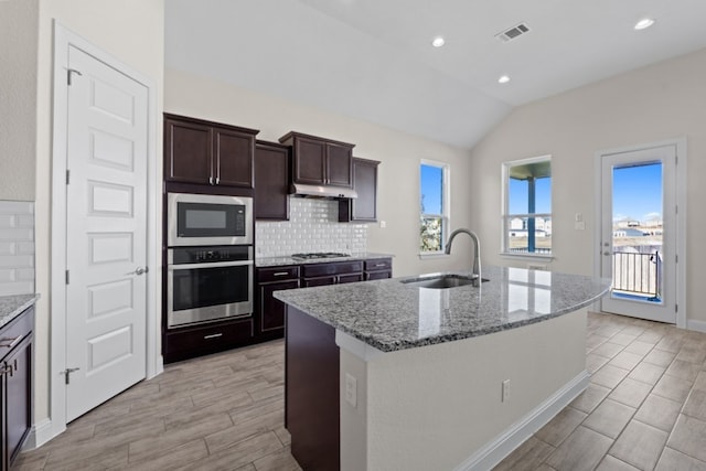 kitchen with light stone countertops, appliances with stainless steel finishes, vaulted ceiling, sink, and an island with sink