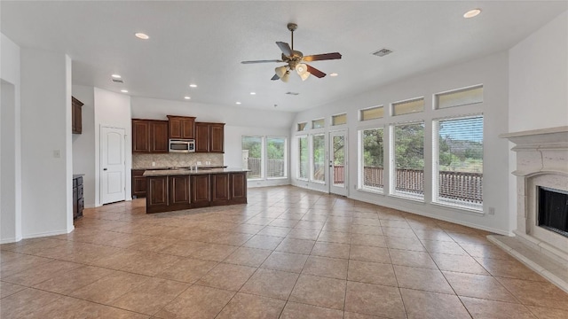 kitchen featuring backsplash, ceiling fan, a center island with sink, a fireplace, and light tile patterned flooring