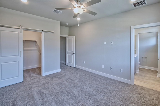 unfurnished bedroom featuring connected bathroom, a walk in closet, light colored carpet, ceiling fan, and a barn door