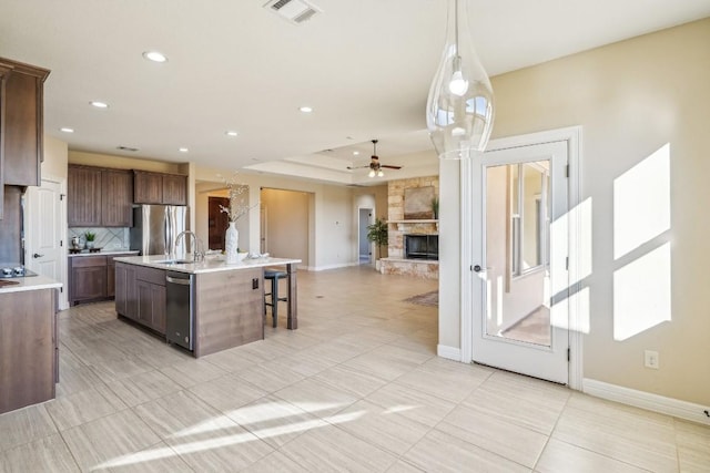 kitchen with a stone fireplace, ceiling fan, an island with sink, appliances with stainless steel finishes, and decorative light fixtures